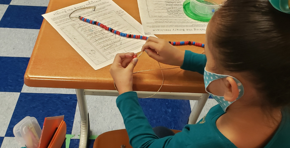 Syracuse Academy of Science first grade Atoms create a beaded Binary necklace. The beads on the necklace correspond to numbers in Binary Code and the alphabet. 